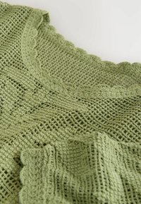 Erkkaanvõtte helerohelisest heegeldatud kangast, mis näitab üksikasjalikke mustreid ja lainelisi ääriseid tekstuurse tekstiilipinna taustal.