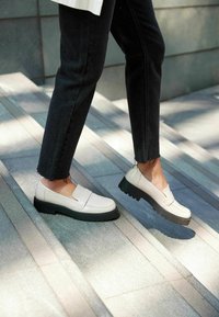 Zapatos slip-on de cuero blanco con una suela negra gruesa, que presentan un diseño plano y un mínimo detalle, usados en escalones de piedra gris.