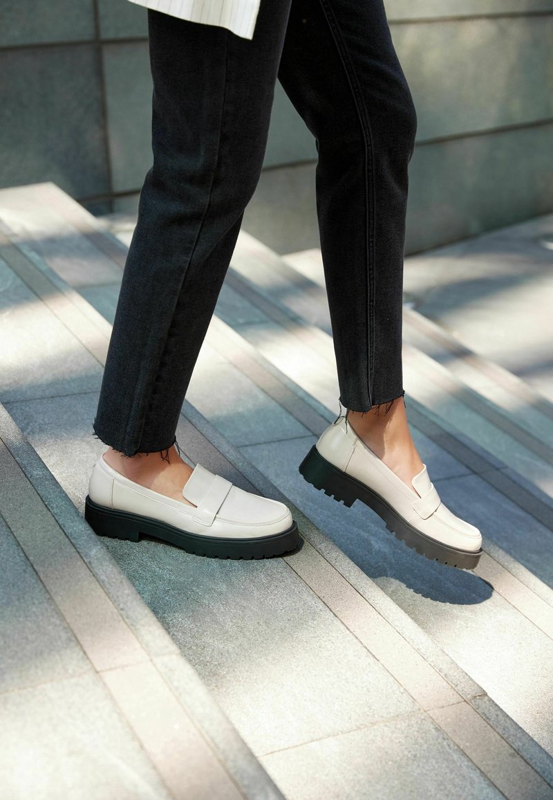 Białe skórzane buty typu slip-on z czarną, masywną podeszwą, o płaskim designie i minimalnych detalach, noszone na szarych kamiennych schodach.