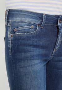 Jeans de mezclilla en azul oscuro con un lavado desgastado. Las características incluyen un diseño de cinco bolsillos, costuras en color naranja y remaches metálicos en los bolsillos.