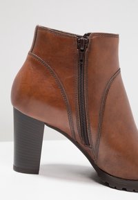 Bota de cuero marrón hasta el tobillo con un tacón alto, que cuenta con un cierre lateral, detalles de costura curvada y una suela de goma texturizada.