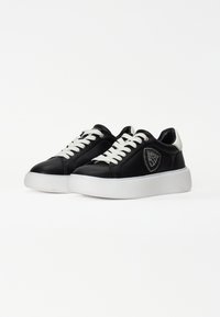 Fekete bőrsneaker fehér fűzővel, texturált fehér talppal és díszes pajzs logóval, csillámtészta díszekkel az oldalon.
