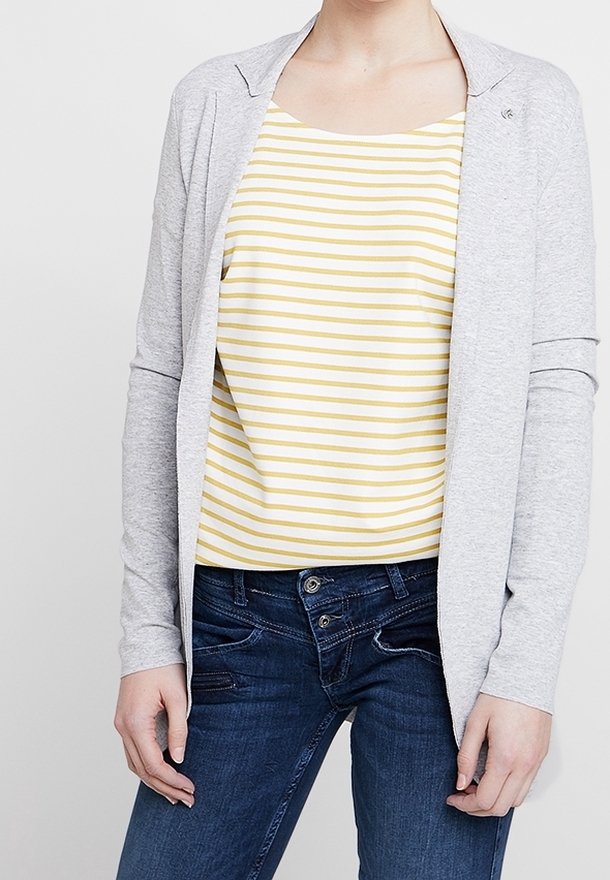 Vaaleanharmaa neuletakki keltaisen ja valkoisen raitaisen hihattoman topin päällä, yhdistetty tummansinisiin farkkuihin. Neuletakissa on hienovarainen kaulussuunnittelu.