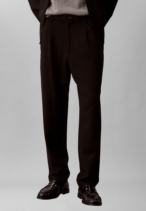 Mann trägt schwarze, maßgeschneiderte Hose mit Knopf- und Reißverschluss, kombiniert mit schwarzen Lederloafers auf grauem Hintergrund.