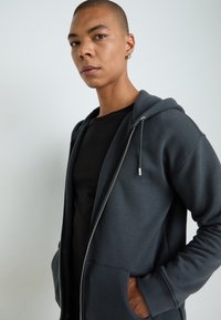 Mørkegrå glidelåshette, med hette, som har en jevn tekstur, sidelommer og metallglidelås, båret over en svart t-skjorte.