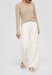Pull beige en tricot avec col en V, associé à un pantalon blanc ample avec des coutures apparentes et des jambes larges, complété par des sandales tressées.