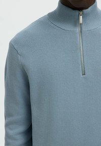 Svijetloplavi pleteni džemper s visokim rebrastim ovratnikom i metalnim zatvaračem na polu, nosi ga osoba.