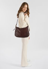 Borsa a spalla in pelle marrone con una morbida texture, forma arrotondata e tracolla regolabile. Caratterizzata da chiusura con zip e minimi dettagli in metallo.