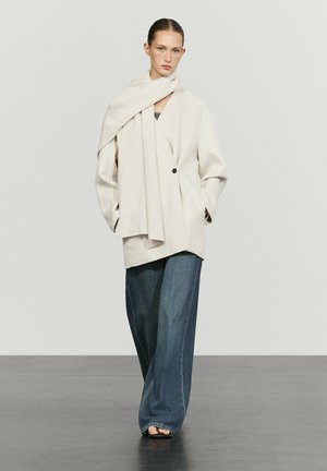 Femme portant un blazer crème surdimensionné avec une écharpe assortie, un jean bleu à jambes larges et des chaussures noires à bouts ouverts, debout devant un fond uni.