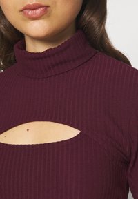 Burgundy ribbade topp med en mock neck och en utskärning nedanför kragen. Tyget har en mjuk textur och en figursydd design.