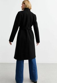 Cappotto di lana nero con vita aderente e cintura, caratterizzato da un colletto alto e spalle strutturate, abbinato a jeans a gamba larga blu.