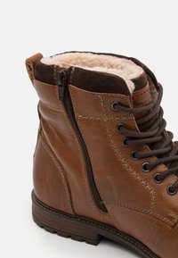 Brune lærstøvler med et robust design, som har glidelås, ullfôr og metalløyler for snøring. Teksturerte sideaksenter er synlige.