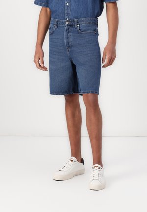 Mężczyzna ubrany w niebieskie szorty dżinsowe, pasującą niebieską koszulę z krótkim rękawem oraz białe trampki, stoi na tle białej ściany.