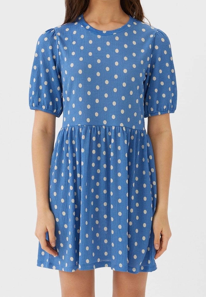Femme portant une robe bleue à manches courtes avec des pois blancs et une taille froncée, debout devant un fond blanc uni.