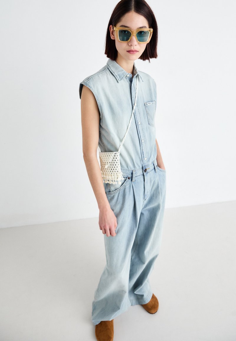 Hellblaue, ärmellose Bluse aus Denim, weit geschnittene Hosen aus hellblauem Denim, braune Schuhe und eine beige geflochtene Umhängetasche. Die Accessoires umfassen große, getönte Sonnenbrillen.