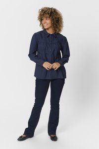 Navyblaues Hemd mit Knopfleiste und Faltendesign, langen Ärmeln und rundem Kragen, kombiniert mit dunkelblauen Schlagjeans und schwarzen Ballerinas.