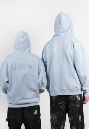 Due persone che indossano felpe con cappuccio azzurro chiaro, con il cappuccio alzato, che mostrano un testo grafico argentato sulle loro schiene, abbinate a pantaloni neri con toppe.
