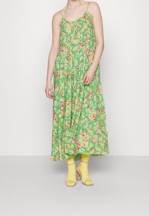 Femme portant une robe maxi sans manches fleurie verte associée à des chaussettes jaunes et des talons ouverts beiges, debout sur un fond blanc.