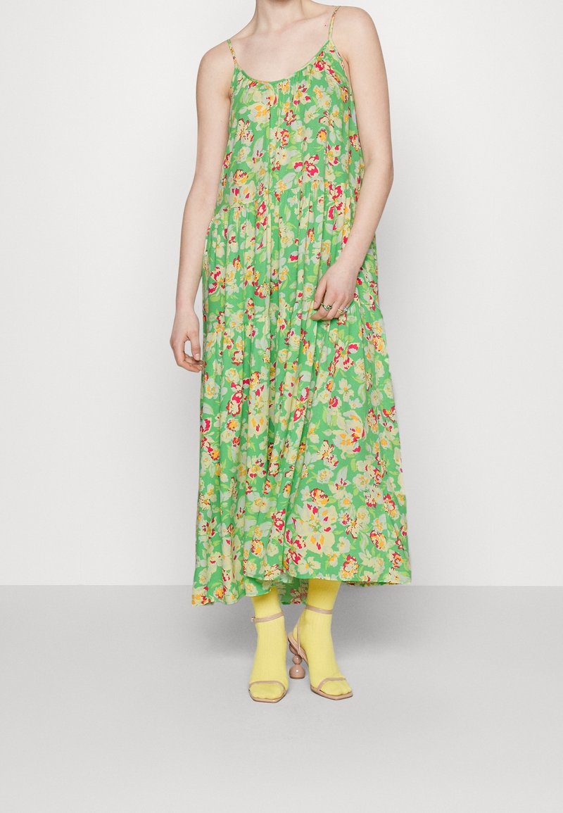 Femme portant une robe maxi sans manches fleurie verte associée à des chaussettes jaunes et des talons ouverts beiges, debout sur un fond blanc.