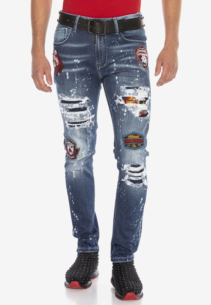 Jean en denim bleu avec des détails déchirés, finition usée, patchs sur les cuisses et taches de peinture blanche. Design ajusté avec passants de ceinture.