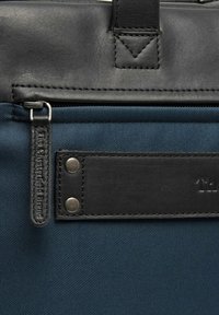 Schwarze Lederakzente auf einer tiefblauen, strukturierten Stofftasche. Verfügt über einen Reißverschluss, ein Markenetikett und Metallnieten auf einem flachen Paneel.