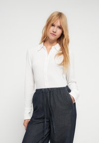 Balti ilgomis rankovėmis marškiniai su sagomis ir apykakle, derinami su tamsiais džinsinio stiliaus kelnėmis, turinčiomis elastingą liemens juostą ir raištelį.