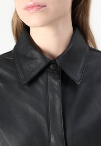 Jachetă din piele neagră, cu guler ascuțit, închidere cu nasturi și o textură netedă. Designul este elegant, cu detalii de cusătură vizibile.