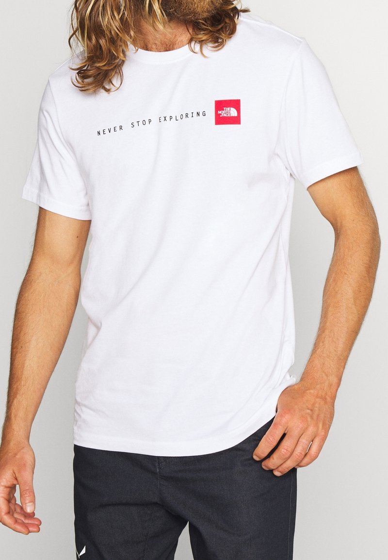 Man med en vit t-shirt med texten "NEVER STOP EXPLORING" och röd The North Face-logotyp, kombinerad med svarta byxor.