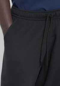 Pantalones deportivos negros con cinturilla elástica, cordón ajustable y bolsillos laterales. Tejido suave con textura lisa y detalles mínimos.