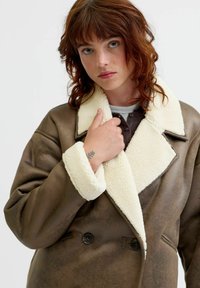 Rudas dirbtinės odos paltas su grietinėlės spalvos avikailio apykakle ir rankogaliais. Yra dvigubo užsegimo sagos ir laisvas siluetas.