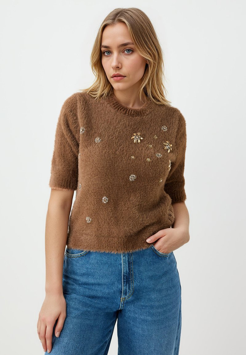 Bruine fluffy trui met korte mouwen, versierd met bloemen. Draagt een blauwe hoge taille jeans, op een lichte achtergrond.