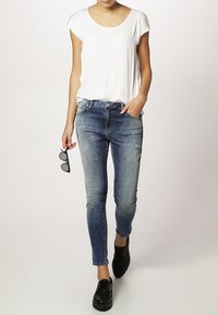 Bílý tričko s krátkým rukávem v kombinaci s tmavě modrými skinny džíny. Černé boty a sluneční brýle drženy v ruce. Jednoduchý, casual design oblečení.