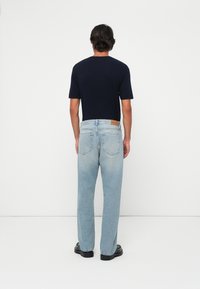 Man wearing black shoes, light blue jeans, and a navy blue short-sleeve shirt standing facing a white wall.
