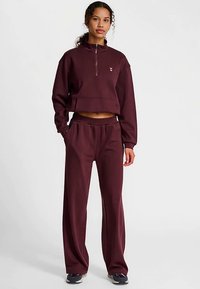 Bordeauxrode tweedelige outfit met een cropped sweater met ritssluiting bij de kraag en wijde broek, gemaakt van zacht materiaal. Heeft minimale branding.