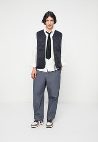 Νεαρός άνδρας στέκεται φορώντας μαύρο πουπουλένιο γιλέκο, λευκό πουκάμισο, μαύρη γραβάτα, χαλαρούς γκρίζους παντελόνες και λευκά αθλητικά παπούτσια, μπροστά από λευκό φόντο.