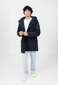 Marine Parka mit Kapuze, Frontreißverschluss und mehreren Taschen. Verfügt über eine glatte Textur und verstellbare Bündchen, kombiniert mit hellen Jeans und Turnschuhen.