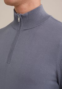 Suéter gris con canalé y cuello alto, que se cierra con cremallera. La tela parece suave y ajustada, con un diseño en capas alrededor del área de la cremallera.