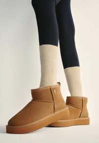 Beżowe zamszowe botki za kostkę na płaskiej gumowej podeszwie. Uzupełnione miękką fakturą i lekko podwyższonym obcasem, w zestawieniu z kremowymi skarpetkami w prążki oraz czarnymi legginsami.