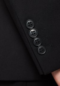 Czarna dopasowana marynarka z rękawem ozdobionym czterema czarnymi guzikami z czterema dziurkami każdy, z wysmakowanymi stempelkami i gładką, strukturalną teksturą.