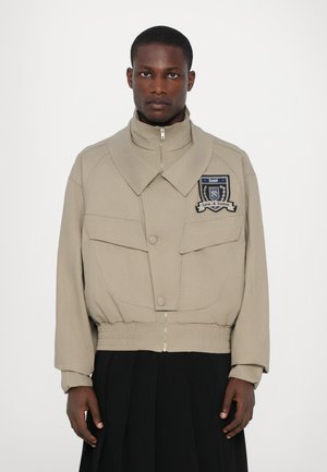 Beige jacket with oversized collar, front pockets, and a zip closure. Features an embroidered patch on the chest. Elastic waistband at the hem.