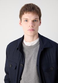 Nehézszövetű, szürke crewneck pulóver felett viselt, galléros nyakú, sötétkék gombos dzseki. Az anyag textúrált hatású, és elöl mellzsebek találhatók rajta.