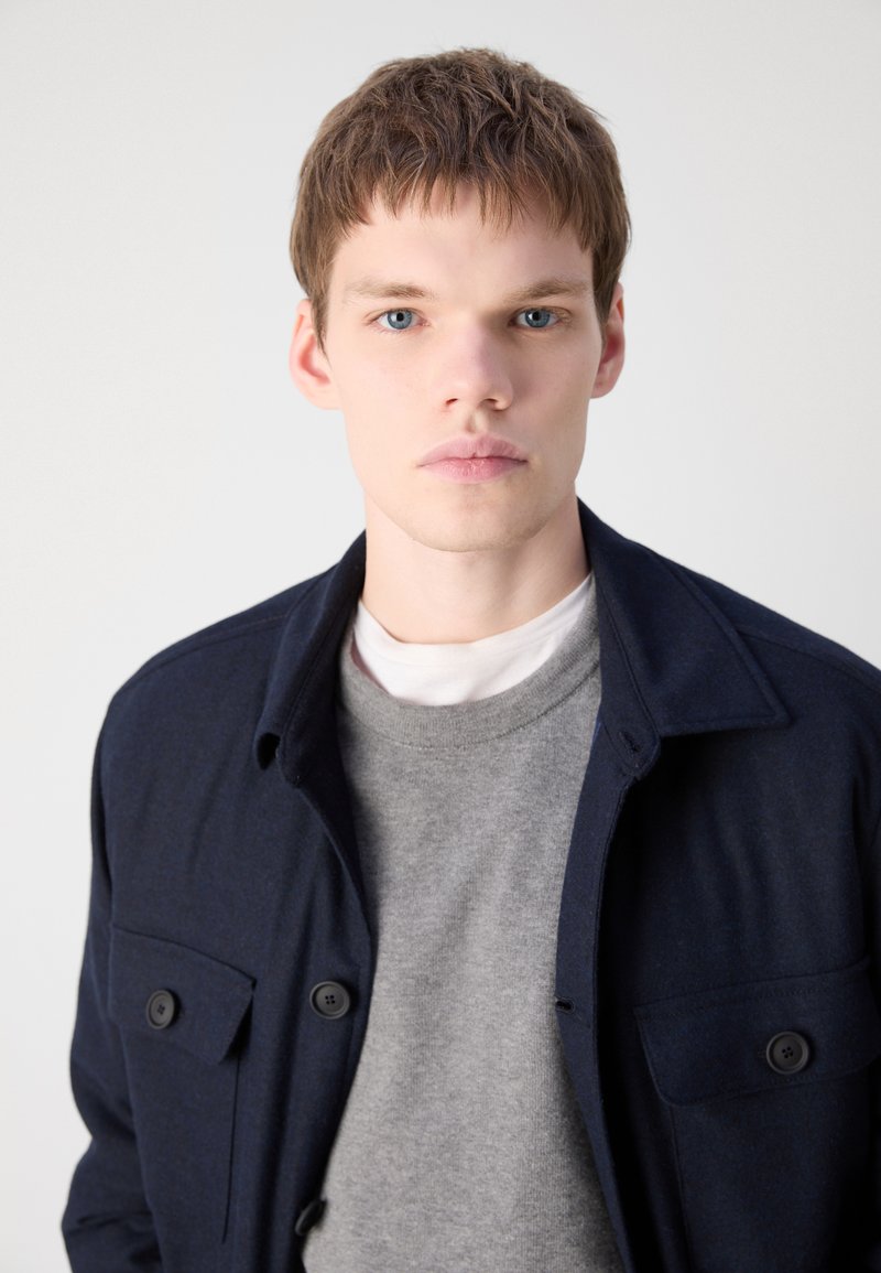 Veste boutonnée bleu marine avec col, portée sur un pull gris à col ras du cou. Le tissu semble texturé et présente des poches poitrine à l'avant.