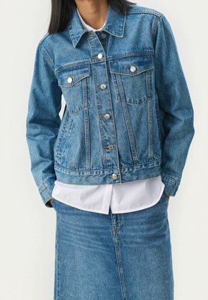 Person trägt eine blaue Jeansjacke über einem weißen Hemd, kombiniert mit einem blauen Jeansrock, die Hände in den Jackentaschen.