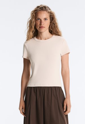 Femme aux longs cheveux blonds ondulés portant un t-shirt uni beige clair à manches courtes et une jupe marron foncé froncée sur fond blanc.