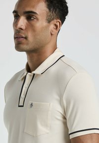 Polo beige avec une finition texturée, présentant une bordure noire sur le col et les manches, et une petite poche avec un logo sur la poitrine.