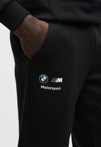 Ruka v kapse černých kalhot s logy BMW a M Motorsport na stehně.