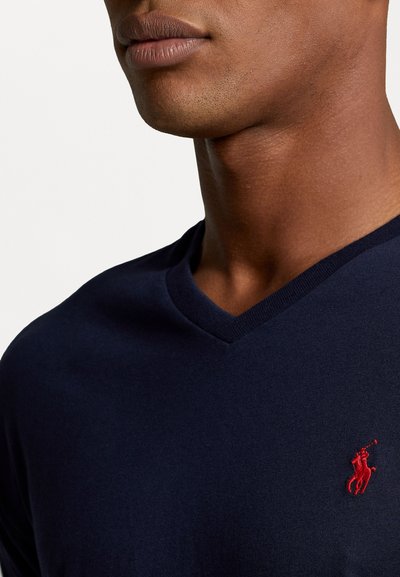 Mörkblå v-ringad t-shirt gjord av mjukt tyg, med en liten röd logotyp på vänster bröst och en slät struktur runt halsringningen.