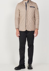 Beige quiltad jacka med upprätt krage och en bröstficka, kombinerad med mörka byxor och svarta skor. Slätt tyg och skräddarsydd passform.