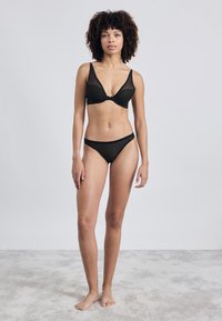 Juodas apatinis komplektas, kuriame yra permatomi puodeliai su plačiomis petnešomis ir atitinkami bikini apatiniai. Medžiaga atrodo glotni su minimalistiniu dizainu.