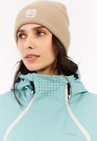 Himmelblaue wasserdichte Jacke mit Belüftungslöchern, kombiniert mit einer hellbraunen gerippten Mütze mit einem Logo-Patch.
